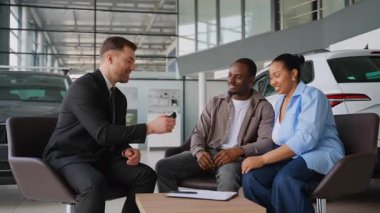 Satış danışmanı neşeli Afrikalı Amerikalı çiftin araba anahtarlarını veriyor, lüks modern galeride otomotiv alımını başarıyla kapatıyorlar, aracın kilometre taşını sembolize ediyorlar.