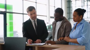 Profesyonel araba satıcısı, modern bayilik galerisinde otomotiv satışlarının başarılı olduğunu gösteren araç satın alma sözleşmesi imzalayan Afro-Amerikan çiftlere rehberlik ediyor.