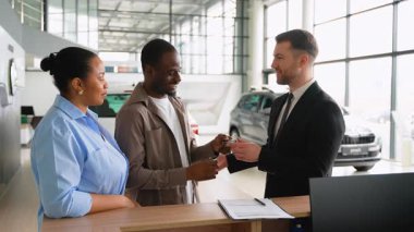 Heyecanlı Afro-Amerikan çifti bir bayiden yeni bir araba alıyor, bir satıcıdan anahtar alıyor ve birbirlerine sarılıyorlar, satın aldıkları için mutlular.