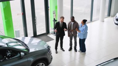 Modern bayilikte Afro-Amerikan çifte yeni otomobil özelliklerini sunan profesyonel araba satıcısı, satın alma seçeneklerini tartışıyor ve yardım sağlıyor.