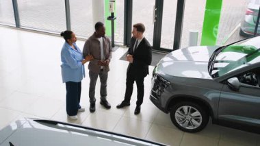 Araç satışını tamamlayan profesyonel satıcı, modern bayilik galerisinde kendine güveni olan Afrikalı Amerikalı çifti selamlıyor.
