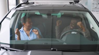 Heyecanlı Afrikalı Amerikalı çift yeni bir arabayı galeride test ediyor, olumlu duygular gösteriyorlar, satın alma kararı verdikten sonra birbirlerine sarılıyorlar.