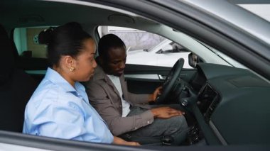 Satış görevlisi, Afro-Amerikalı çifte bir bayide yeni bir arabanın özelliklerini keşfetmede yardım ediyor, güneşliği ayarlıyor ve gösterge panelini kontrol ediyor.