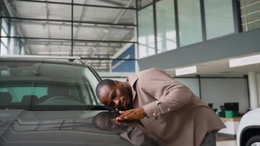 Hevesli bir adam modern, geniş bir otomobil galerisinin içinde parlak, koyu renkli bir arabanın kaputuna nazikçe dokunuyor. Potansiyel alışverişe olan hayranlığını ve heyecanını dile getiriyor.