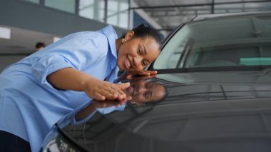 Mutlu Afro-Amerikalı kadın bir galeride yeni bir arabanın kaputunu nazikçe okşuyor, alışverişi tamamlamadan önceki anın tadını çıkarıyor.