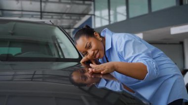 Mutlu Afrikalı Amerikalı kadın yeni arabasının kaputuna nazikçe dokunuyor. Pürüzsüz çizgilerine ve araba galerisindeki kusursuz boyasına hayran kalıyor.