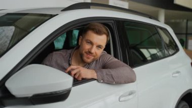 Yeni alınmış bir araçta oturan adam, elinde heyecanlı bir ifadeyle anahtarlar, galeride otomotiv alımını kutluyor.