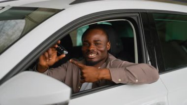 Araba sahibi, gülümseyen bir gülümsemeyle anahtarlarını sergiliyor, modern galeride başarılı araba satın alımını kutluyor, yeni otomobil sahibi olmanın sevincini ve gururunu dile getiriyor.