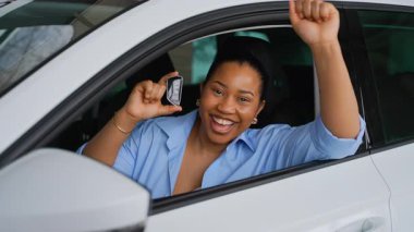 Mutlu Afrikalı Amerikalı kadın yeni arabanın sürücü koltuğunda oturuyor, anahtarları gösteriyor ve kameraya gülümsüyor, araba galerisinde alışverişi kutluyor.