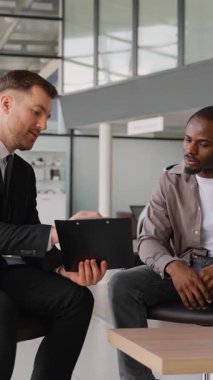 Profesyonel araba satıcısı, Afrika kökenli Amerikalı bir çiftin modern galeri sergi salonundaki yeni araçlarını seçmelerine yardım ederken sözleşmeyi sunuyor.