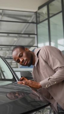 Mutlu Afro-Amerikan bir adam otomobil galerisinde yeni bir arabanın kaputunu nazikçe okşuyor, alışverişi sonuçlandırmadan önceki anın tadını çıkarıyor.