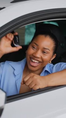 Araba galerisindeki yeni arabasında oturan Afrikalı Amerikalı kadın, elinde yeni alınmış bir anahtar ve başarılı bir otomotiv alımından yayılan mutluluk var.
