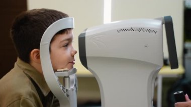 Genç çocuk sabırla göz muayenesinden geçiyor, kafası özel bir cihaza yerleştirilirken, görünmeyen bir göz doktoru gelişmiş ekipmanları kullanıyor.