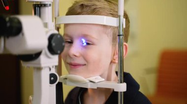 Pediatrik göz doktoru modern bir yarık lamba biyomikroskobuyla çocuğun gözünü inceliyor ve hastaların gözbebeklerine mavi ışık ışını yansıtıyor.