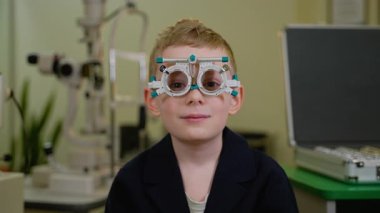 Genç çocuk göz doktorunun ofisinde sabırla oturuyor, kapsamlı bir göz muayenesi sırasında bir deneme çerçevesi takıyor, doktor görüşünü değerlendirirken yüz ifadeleri hafifçe değişiyor.