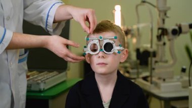 Deneyimli göz doktoru, uzman klinik ortamında detaylı tıbbi görüş kontrolü sırasında profesyonel deneme çerçevesi kullanan genç bir çocuk üzerinde kapsamlı göz muayenesi yapıyor.
