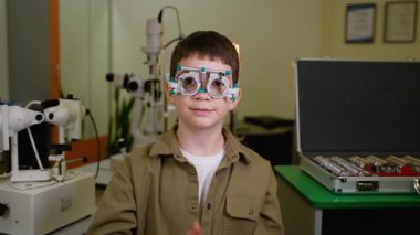Deney çerçevesi giyen neşeli pediatrik hasta, tıbbi klinikteki destekleyici göz doktoruyla kapsamlı göz muayenesi sırasında kendinden emin bir şekilde onaylıyor.