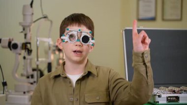 Kapsamlı göz muayenesi sırasında deneme çerçevesi takan genç hasta, pediatrik görüş taraması sırasında göz doktorunun ofisindeki farklı görme alanlarını işaret ediyor.