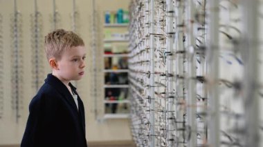 Genç sarışın çocuk optik mağaza rafından şık göz giysileri seçiyor, çerçeveleri dikkatlice deniyor ve görüş düzeltmesi için alışveriş yaparken aynada görüntüyü kontrol ediyor.