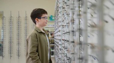 Yeni takılmış gözlük takan neşeli genç bir çocuk. Optik çerçevelerin duvar ekranının yanında duruyor. Kameranın lensine bakarken neşe saçıyor.