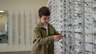 Genç bir çocuk bir optik mağazada çeşitli gözlükler seçiyor ve deniyor. Gelişmiş görme yeteneği için mükemmel bir uyum sağlamak için farklı çerçeveler keşfediyor.