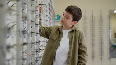Genç çocuk optik mağaza çerçevelerini karıştırıyor, farklı tarzları hevesle denerken dikkatlice gözlük seçiyor, mükemmel görme aksesuarı için birden fazla seçeneği inceliyor