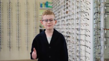 Neşeli genç çocuk gururla yeni reçeteli gözlük takıyor, modern optik dükkanında dikilirken başparmak hareketi yapıyor, gelişmiş görme ve şık gözlüklerle tatmin olduğunu ifade ediyor.