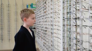 Genç çocuk şık gözlükleri dikkatlice seçiyor, farklı çerçeveleri karşılaştırıyor ve onları optik vitrin görüntülerini incelerken deniyor.