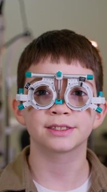 Geniş kapsamlı göz muayenesi sırasında deneme çerçevesi takan genç hasta, pediatrik göz hastalıkları kliniğinde tedavi görürken güvenle gülümsüyor.