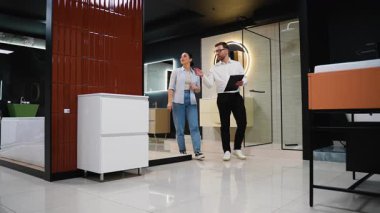 Modern bir galerideki bir kadına banyo malzemeleri gösteren profesyonel bir danışman. Seçenekleri tartışıyor ve yenileme projesi için uzman tavsiyesi veriyor.