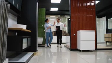 Modern bir sergi salonundaki bir çifte banyo armatürleri, fayanslar ve tesisat seçeneklerini gösteren profesyonel bir danışman. Ev yenileme projeleri için bilgilendirilmiş kararlar almalarına yardımcı oluyor.