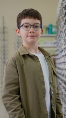 Yeni gözlük takan genç çocuk, parlak optik mağaza ortamında dikilirken gelişmiş görüşle tatmin olduğunu belirterek kendinden emin bir şekilde baş parmak hareketi yapıyor.