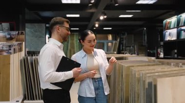 Modern bir sergi salonunda fayans seçen bir kadına yardım eden profesyonel danışman, uzman tavsiyeleri sunuyor ve ev yenileme projesi için çeşitli seçenekleri tartışıyor