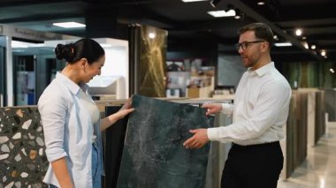 Modern bir sergi salonundaki müşteriye farklı fayans örnekleri gösteren satış asistanı, çeşitli iç tasarım projeleri için doku, renk ve uygunluğu tartışır.