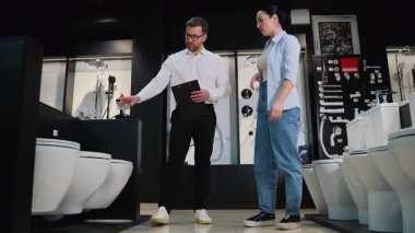 Tuvalet danışmanı, tuvaletteki mobilya mağazasında çalışan bir kadına farklı tuvalet modelleri gösteriyor, özellikleri tartışıyor ve ev yenileme projesi için seçim yapmasına yardım ediyor.
