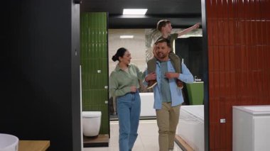 Aile, banyo fayanslarını birlikte seçiyor, baba, oğlunu omuzlarında taşırken modern sergi salonundaki satış elemanına danışıyor, ev yenileme sürecini ortaklaşa sergiliyor.