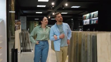 Genç bir aile daire tadilatı için fayans seçiyor, nalbur dükkanında danışmanla yürüyor, banyo veya mutfak için malzeme tartışıyor ve seçiyor.