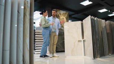 Modern sergi salonu düzeninde ev yenileme projesi için çeşitli zemin seçeneklerini araştırarak genç çifte fayans seçim sürecinde rehberlik eden profesyonel iç mimari danışmanı
