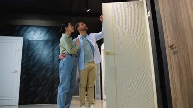 Genç çift çeşitli iç kapı modellerini inceliyor, modern bir sergi ortamında bir satış danışmanından rehberlik alıyor, stilleri ve özellikleri tartışıyor.