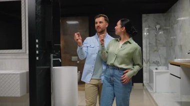 Genç çift profesyonel tasarımcıyla görüşüyor, banyo armatürlerini dikkatle inceliyor ve şık, çağdaş sergi salonu ortamında yenileme seçeneklerini tartışıyor.