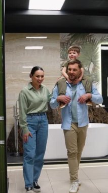 Genç aile profesyonel danışman ile banyo tasarım seçeneklerini araştırıyor, çocuk babasının omuzlarına tünemiş, gösterişli bir sergi salonunda iş birliği yapıyor.