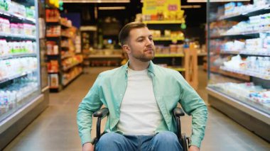 Erkek müşteri bakkalda bağımsız olarak geziniyor, tekerlekli sandalyeden ürünleri seçerken gülümsüyor, alışveriş deneyimlerinde güven ve neşe ifade ediyor