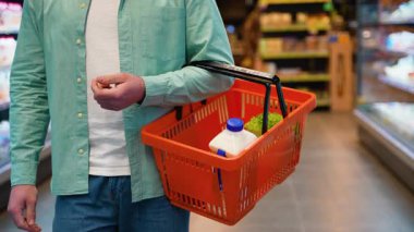 Kendine güveni tam bir erkek alışverişçi, marketin koridorlarında geziniyor, turuncu sepetle taze süt ve marul taşırken sağlıklı gıda ürünleri seçiyor.