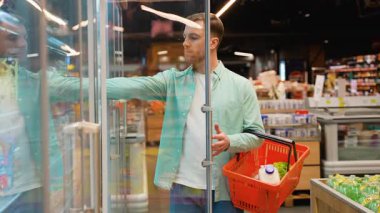 Genç müşteri marketteki soğutulmuş koridordan taze yumurta seçiyor, elinde süt ve sebze dolu bir alışveriş sepeti tutuyor, son kullanma tarihini dikkatlice kontrol ediyor.