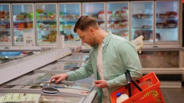 Süpermarket müşterisi dondurulmuş yiyecekleri dondurulmuş bölümden seçiyor, elinde bir alışveriş sepeti tutuyor ve son kullanma tarihlerini ve malzemelerini kontrol ediyor.