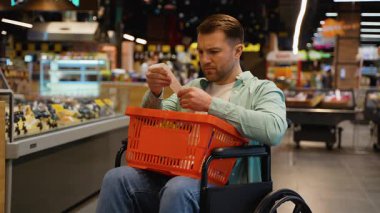 Tekerlekli sandalye kullanıcısı süpermarkette oturuyor, şaşırdığını ve hayal kırıklığını dile getirirken, alışveriş sepetinin yanında duran son derece uzun market fişini inceliyor.