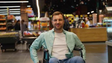 Tekerlekli sandalyede oturan, markette kameraya bakıp gülümseyen, alışverişi seven, bağımsız ve mutlu hisseden kendine güvenen bir müşteri.