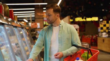 Genç adam soğutulmuş raflardan peynir seçiyor, elinde alışveriş sepeti tutuyor, etiketini okuyor, ürünleri karşılaştırıyor, karar veriyor, sepete peynir koyuyor, süpermarketten alışveriş yapıyor.