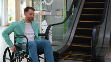 Alışveriş merkezinde yürüyen merdivene bakan tekerlekli sandalye kullanan genç adam, kentsel kamu altyapısındaki hareketlilik sorunları ve seyrüsefer stratejilerini değerlendiriyor