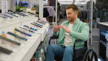 Tekerlekli sandalyedeki mutlu adam gülümsüyor ve akıllı telefon kullanıyor. Teknoloji mağazasında yeni cep telefonunu seçip test ediyor. Elektronik mağazasında alışveriş yapmaktan hoşlanıyor.